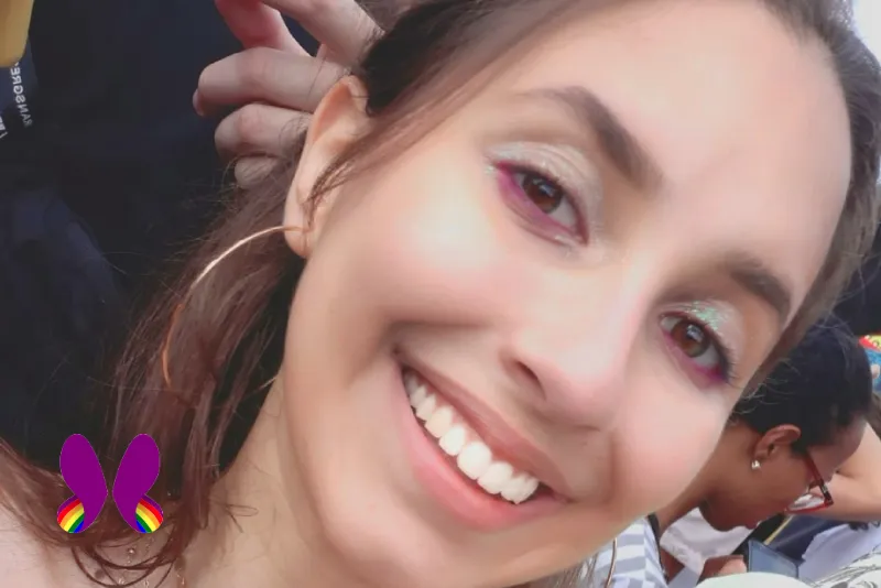 Foto de Laís sorrindo, usando brincos de argola dourados, maquiagem com glitter e cabelos presos na parte de cima. A logo do projeto está no canto inferior esquerdo.