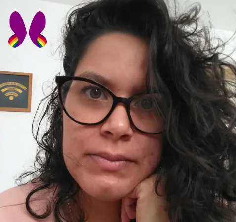 Elayne está com o rosto apoiado na mão, com uma expressão séria. Ela usa óculos, tem cabelos médios escuros, é uma mulher branca.